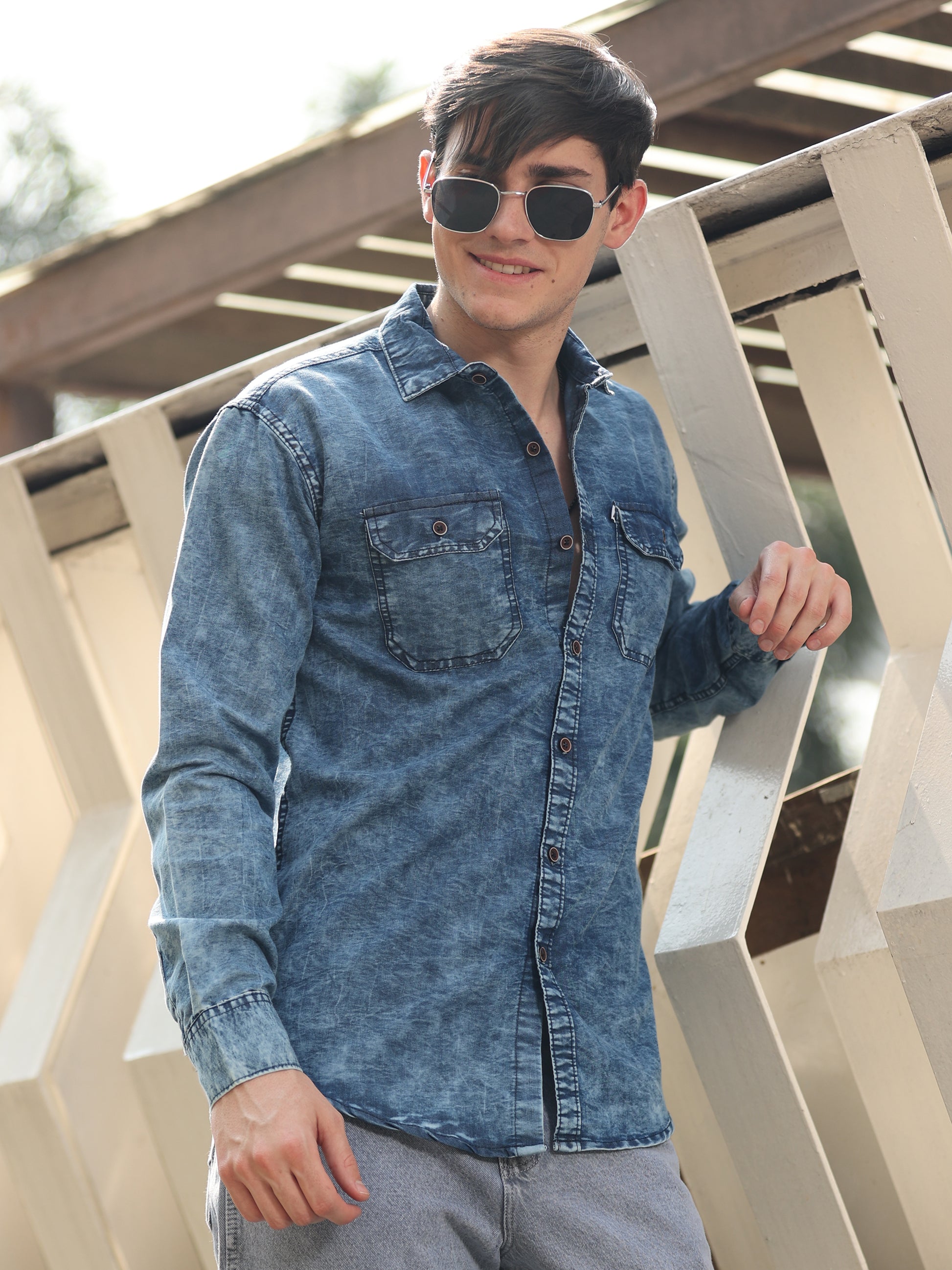 Young man in fashionable blue denim shirt styled with sunglasses and light grey jeans