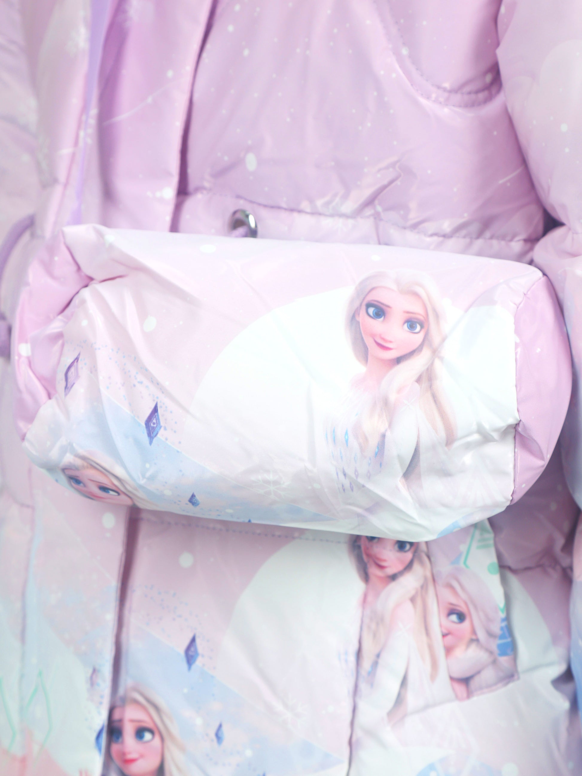 Close-up of a light purple puffer jacket with matching crossbody bag, both featuring printed images of Frozen character Elsa in a snowy, pastel-themed design.
