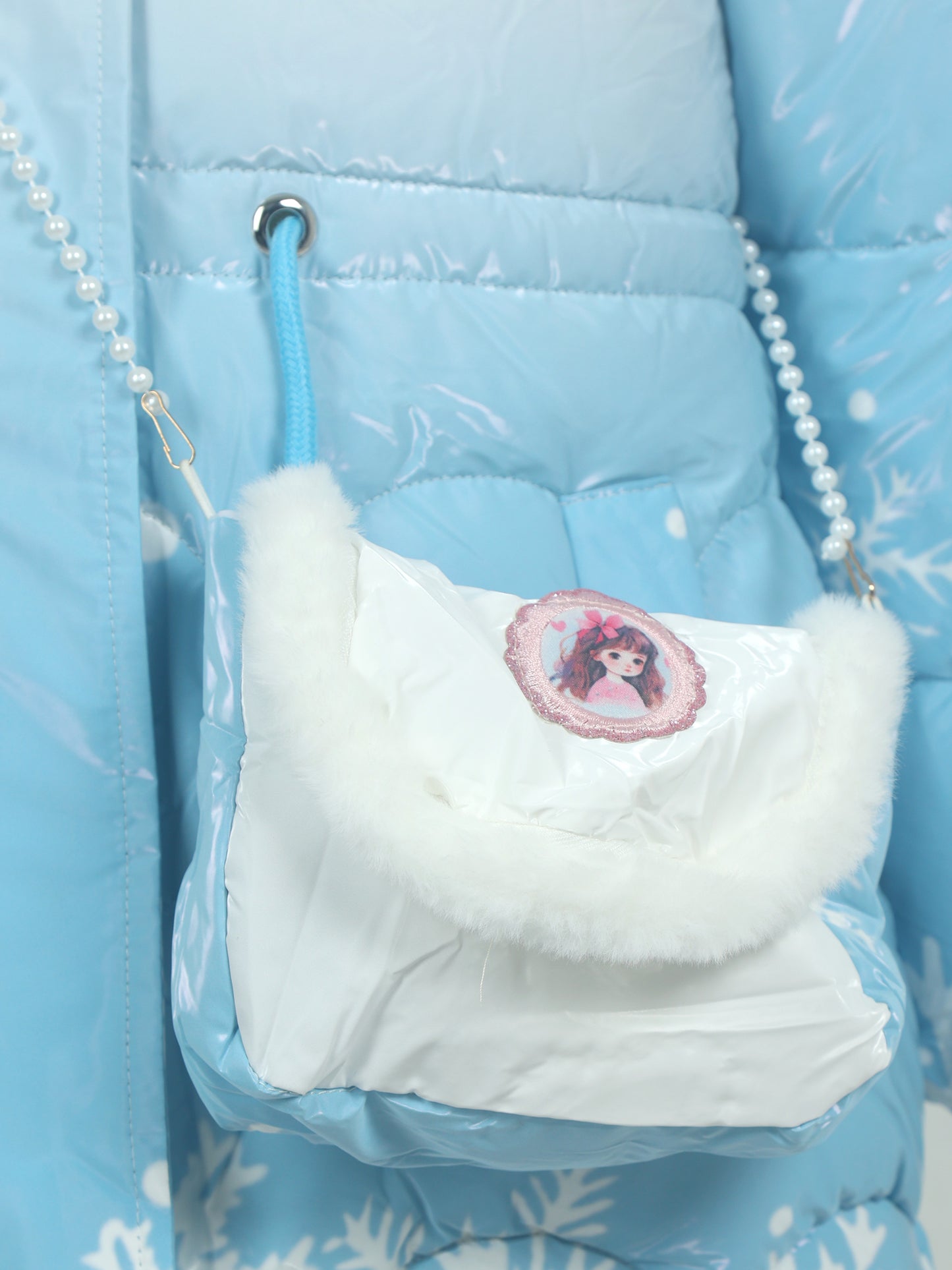 Close-up of a small light blue shoulder bag with a white faux fur flap and pearl strap, featuring an embroidered patch of a doll worn over a matching blue winter jacket with snowflake patterns.