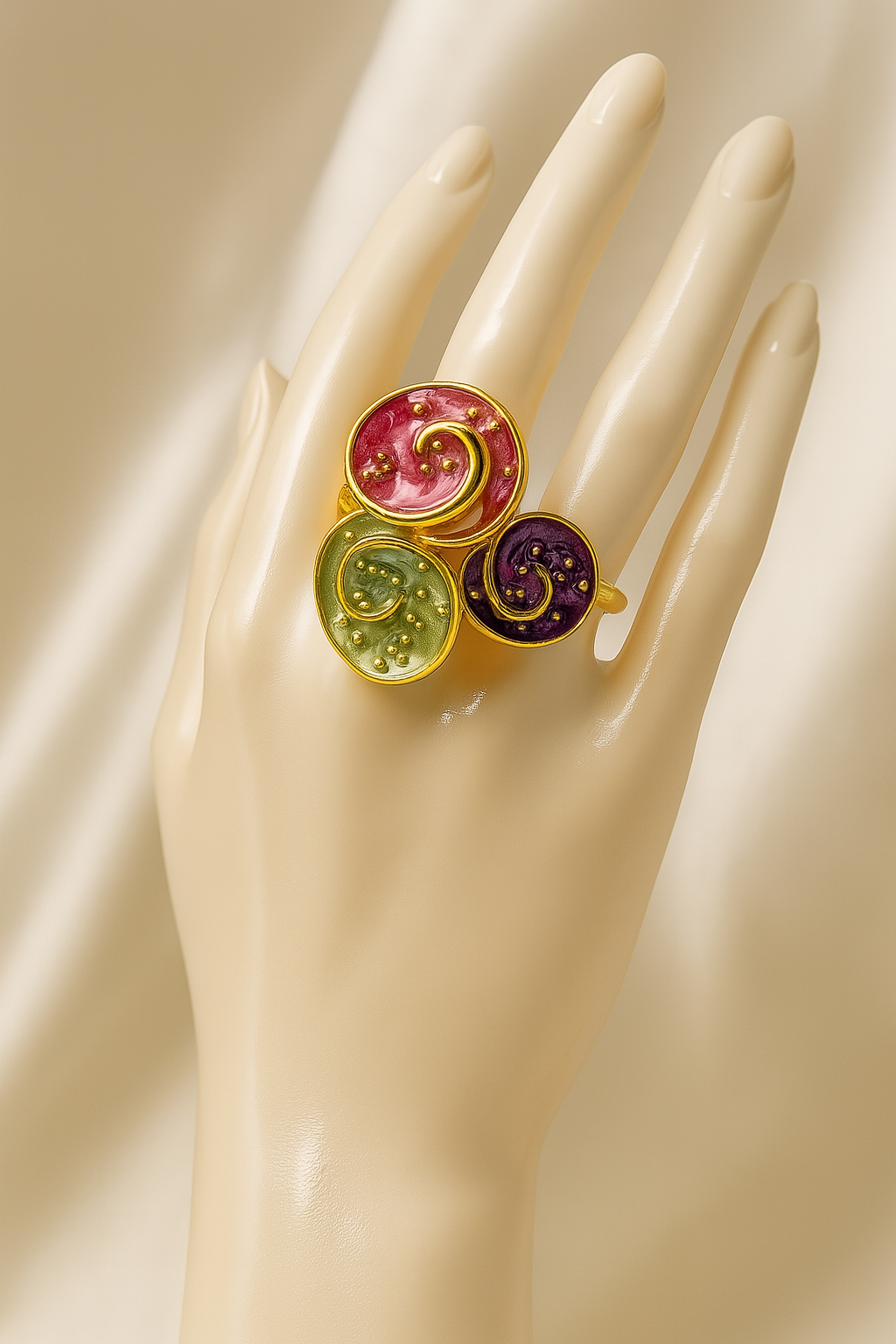 Gold-plated triple spiral statement ring with colorful enamel, worn on a mannequin hand against a soft beige satin background.