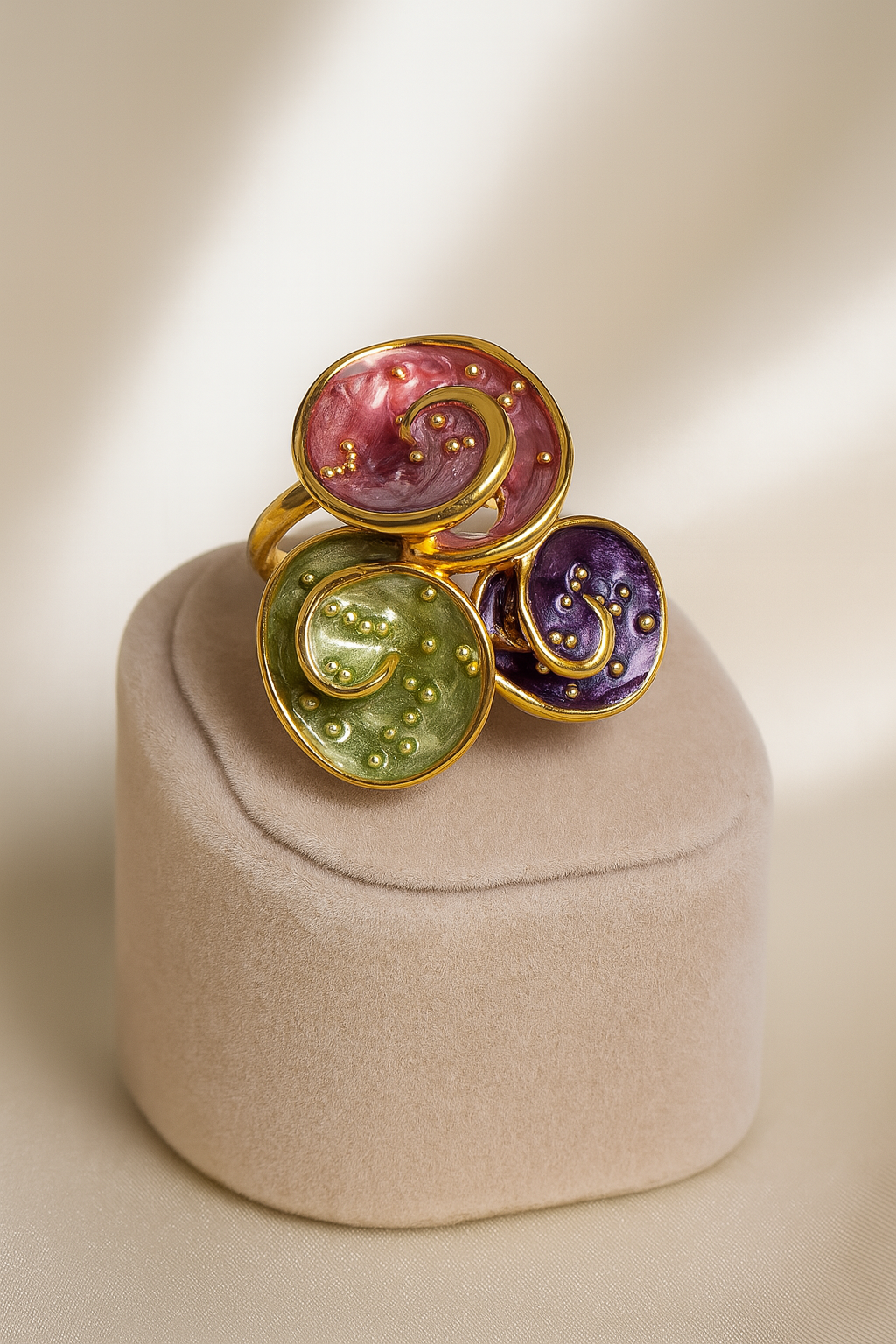 Elegant gold-plated triple spiral enamel ring in red, green, and purple hues, displayed on a beige velvet ring stand with a soft satin backdrop.