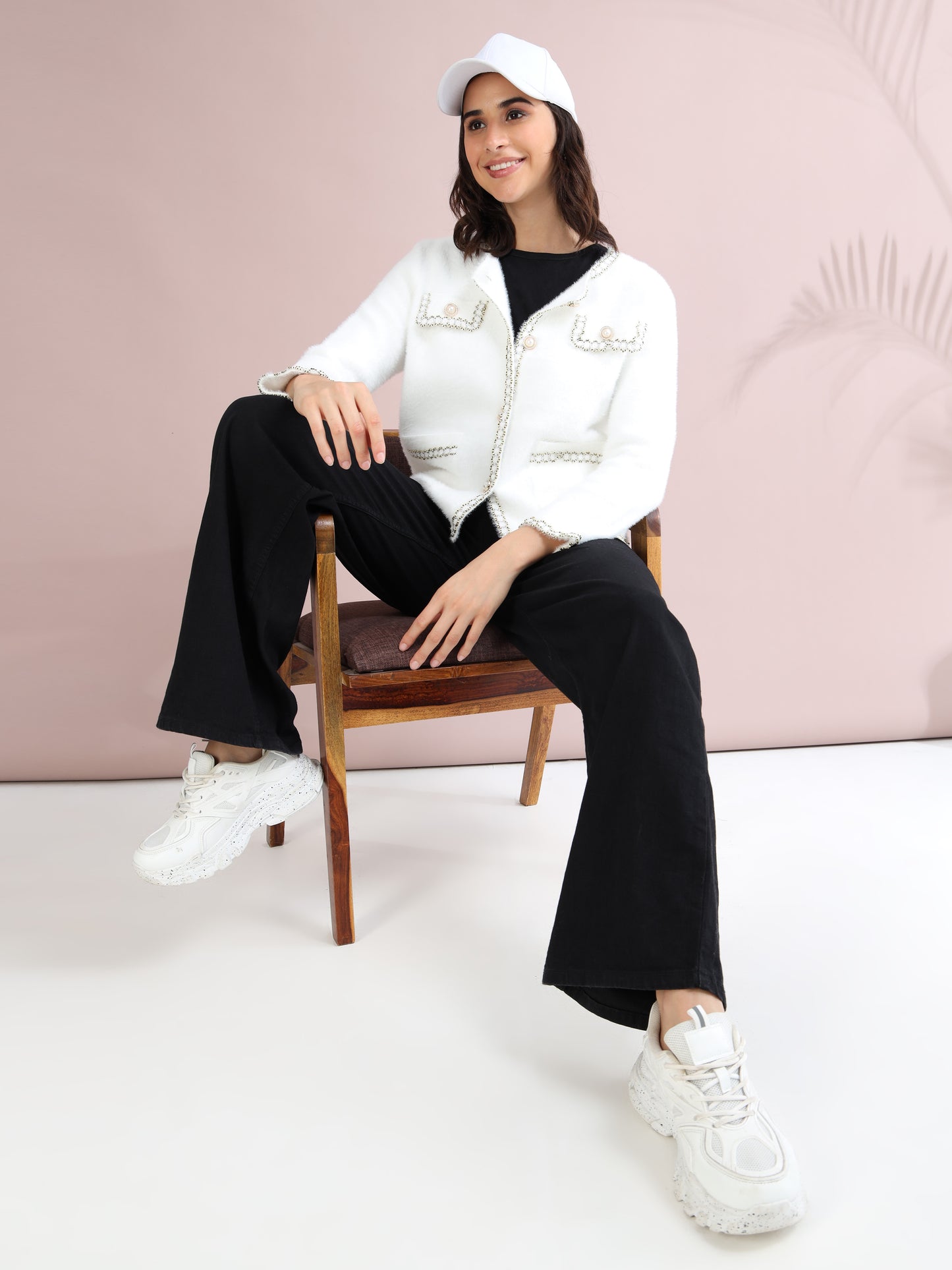 Woman seated wearing white pearl button cardigan, black wide-leg pants, white sneakers, and white cap for a chic casual style.