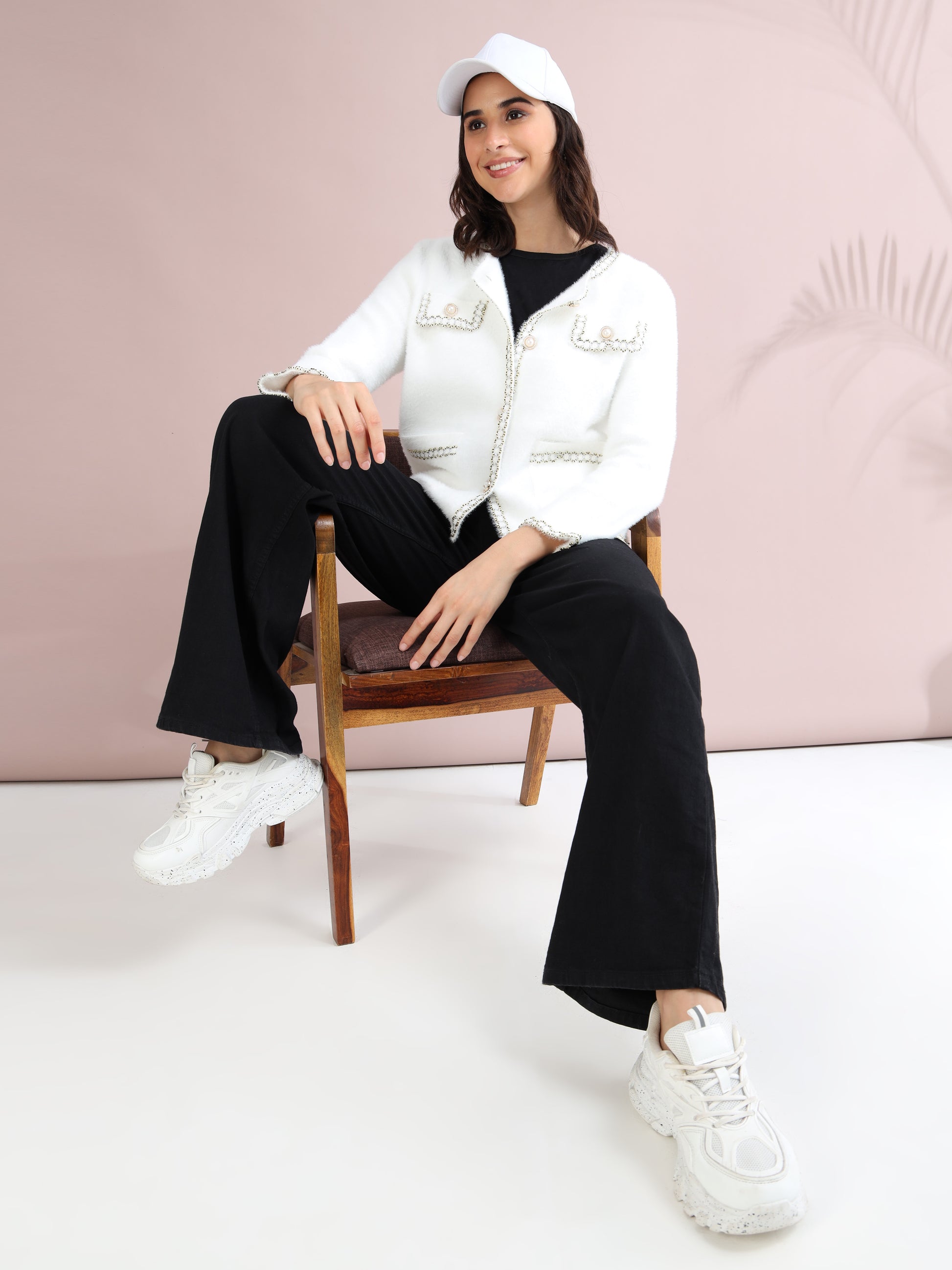 Woman seated wearing white pearl button cardigan, black wide-leg pants, white sneakers, and white cap for a chic casual style.
