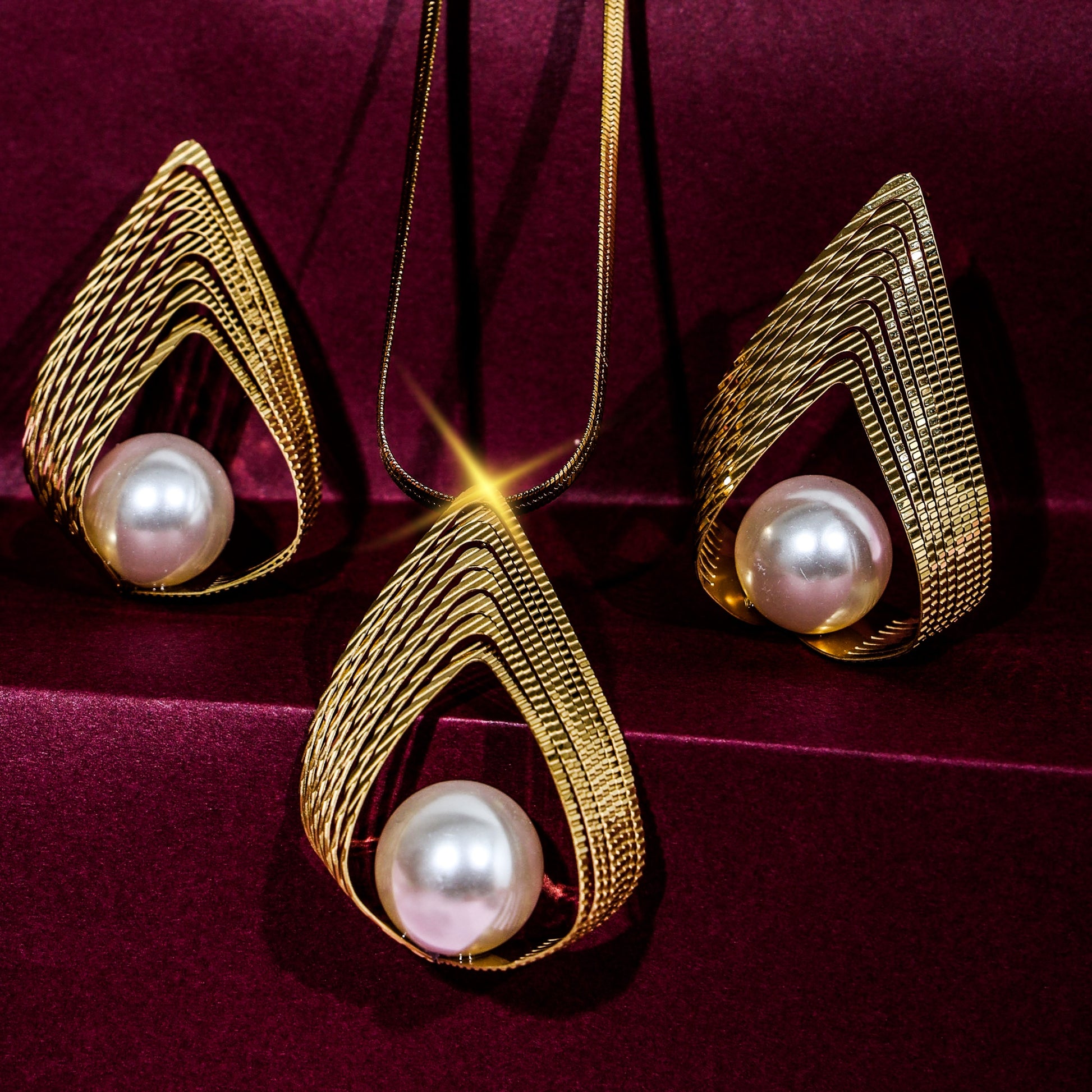 Close-up of a gold-plated teardrop pearl necklace and earrings set on a rich maroon velvet surface with a sparkling highlight effect on the pendant