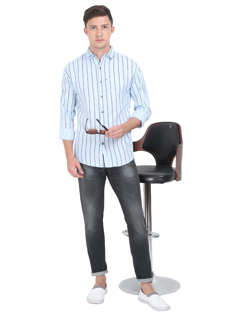 Man wearing a light blue striped shirt and black jeans standing next to a black bar stool on a white background