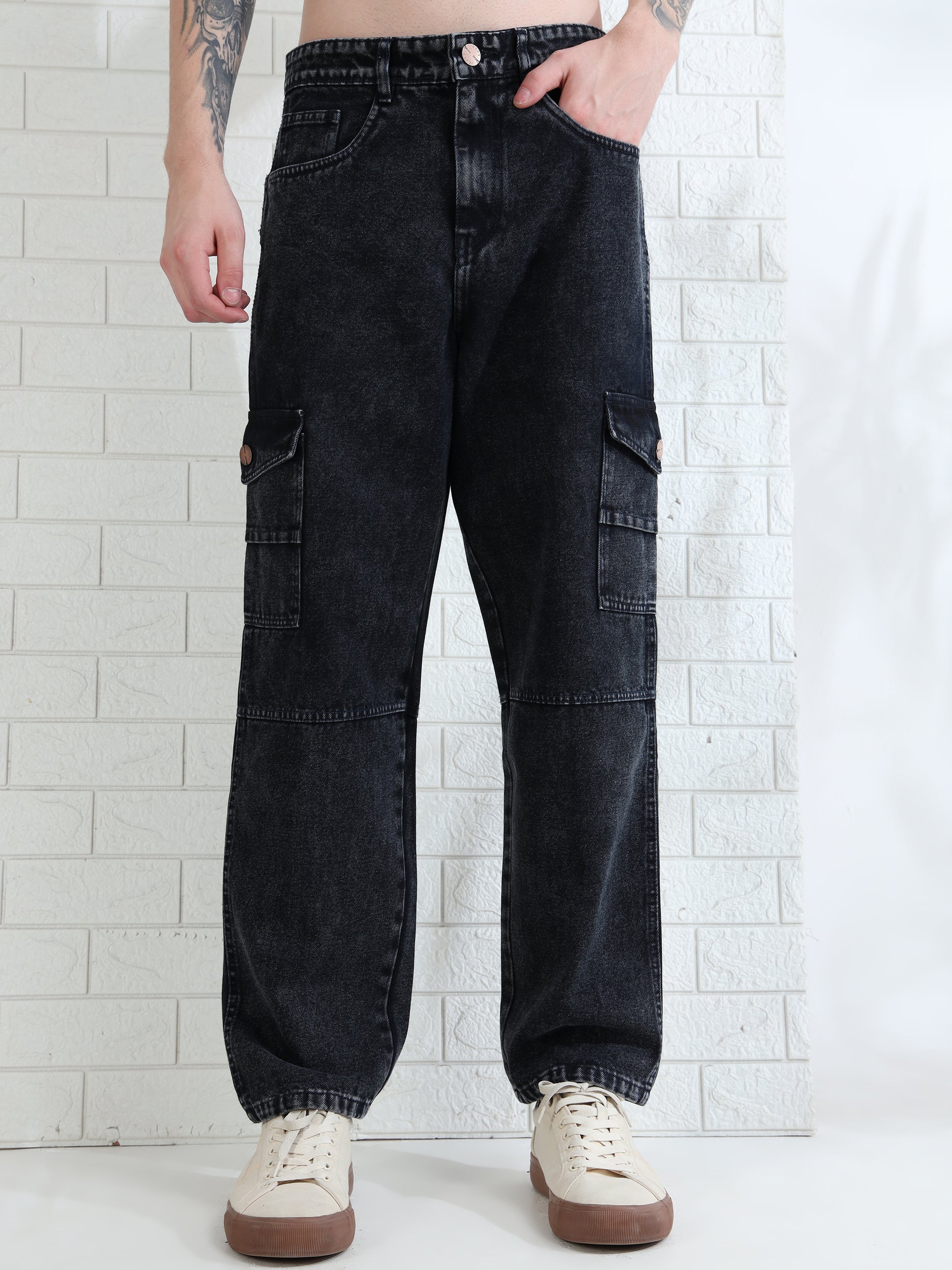 A person standing against a white brick wall wearing black mid-rise denim jeans with multiple utility pockets and white sneakers.