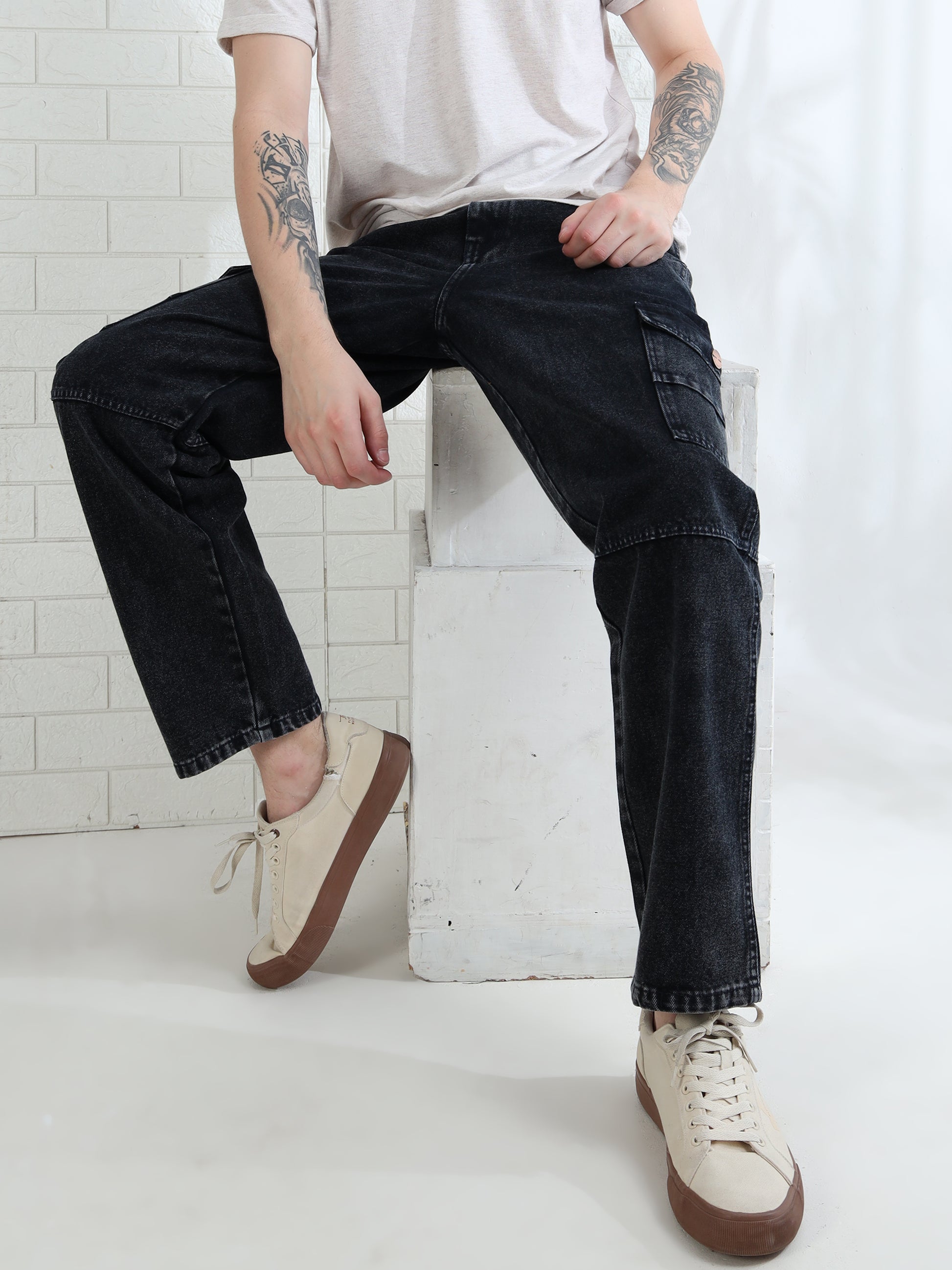 A person sitting on a white cube-shaped bench, wearing mid-rise black denim jeans with multiple utility pockets and a white t-shirt, with their feet resting on the same bench.