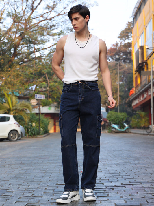 A person wearing a white sleeveless top and dark blue baggy cargo jeans standing on a street.