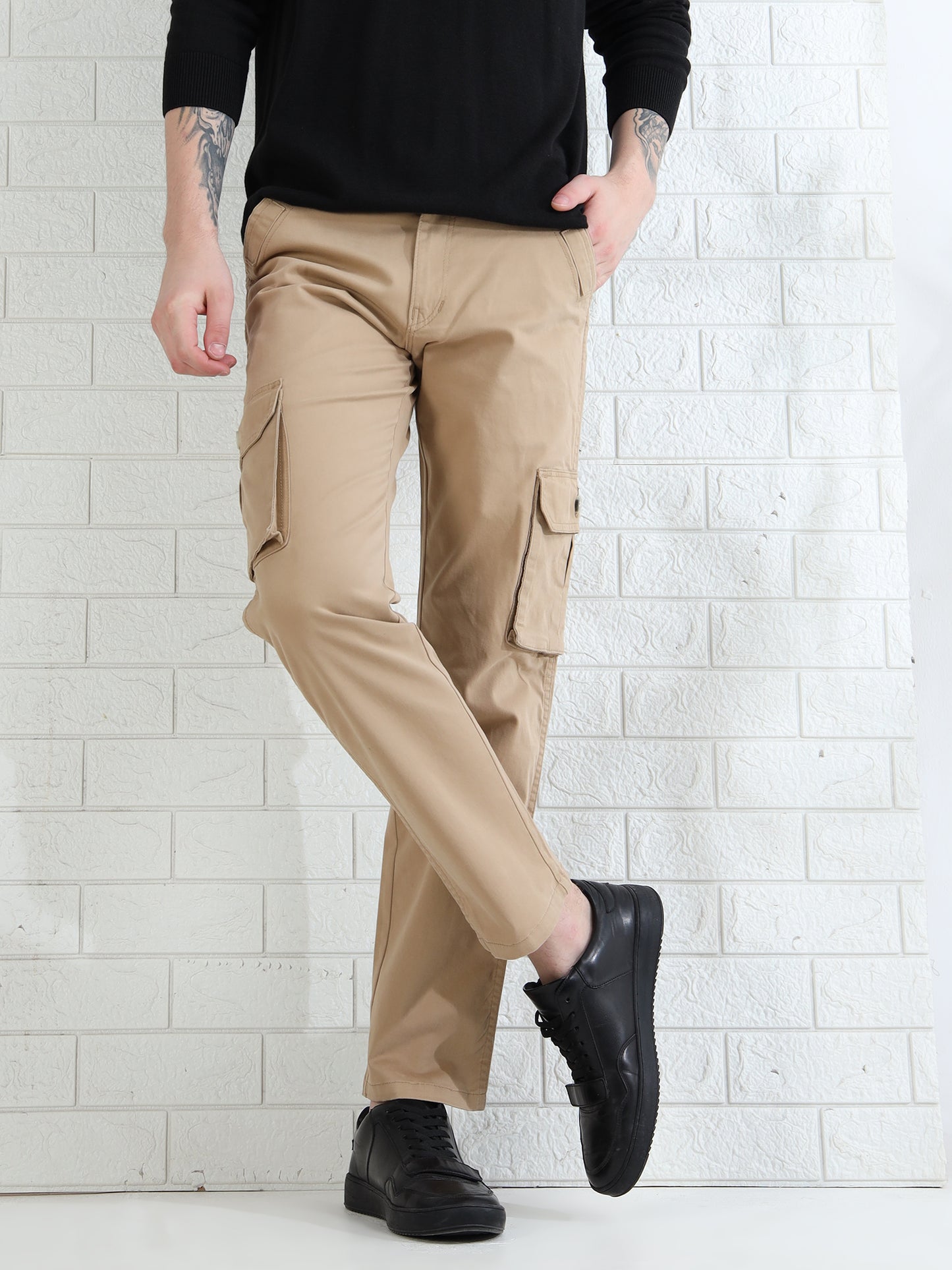 A person standing against a white brick wall, wearing beige cargo pants with multiple pockets and a black top, paired with black shoes.