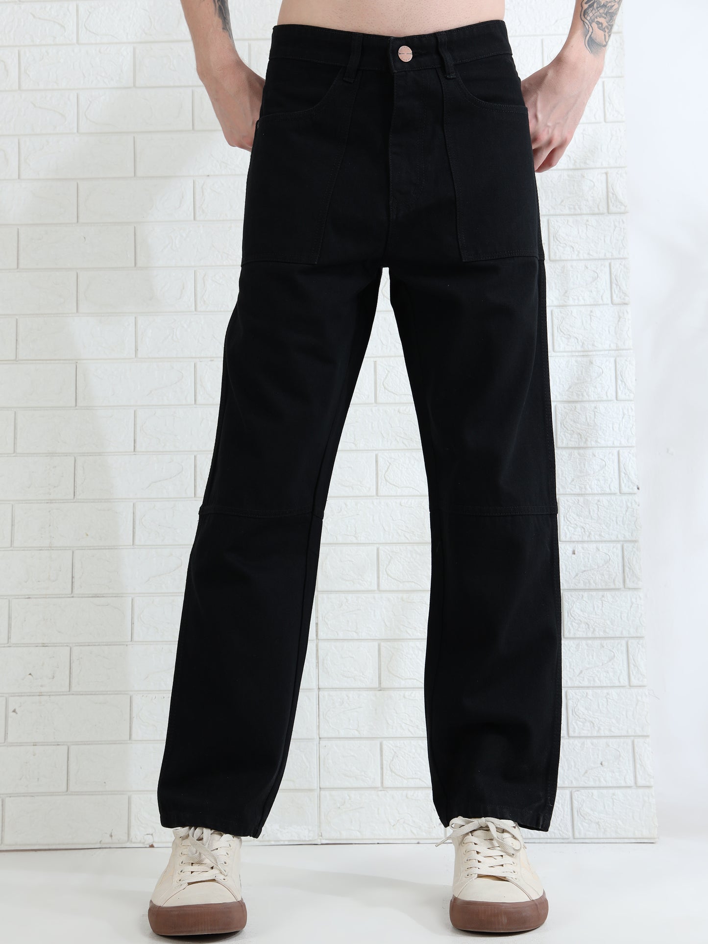 A person standing against a white brick wall, wearing black mid-rise waist denim pants with four pockets and button-closure waistband.