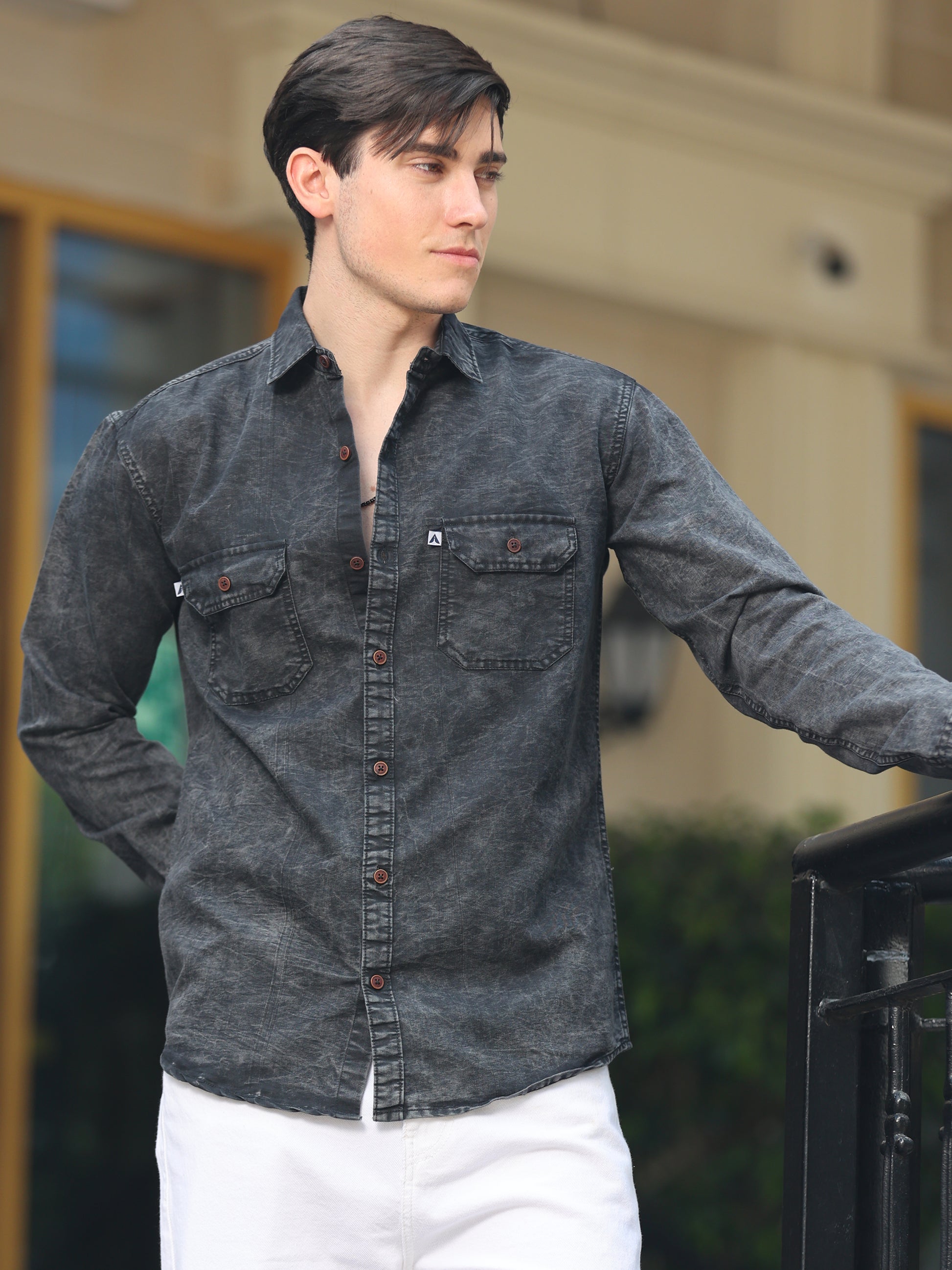 A man wearing a long-sleeved, black denim shirt with a regular collar, buttoned cuffs, and two chest pockets. He is also wearing white pants.