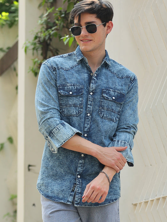 A man wearing a blue denim shirt with full sleeves, a regular collar, and two pockets, paired with light gray trousers and sunglasses.