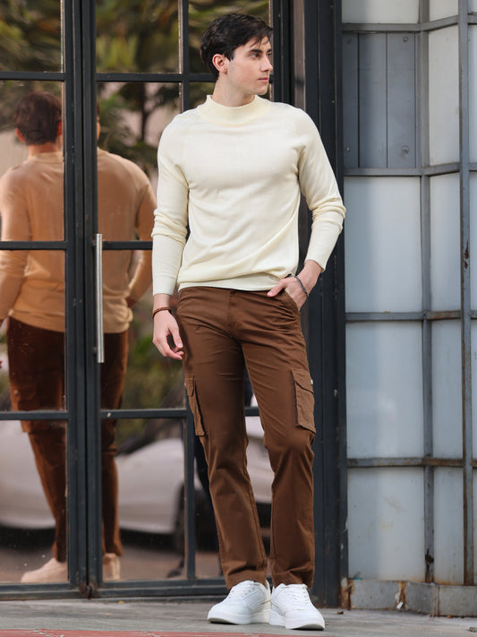 A person wearing brown cargo pants and a beige sweater standing near a glass door.