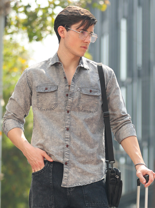 A man wearing a grey denim shirt with a regular collar, full sleeves, and two chest pockets. He is also carrying a bag and wearing glasses.
