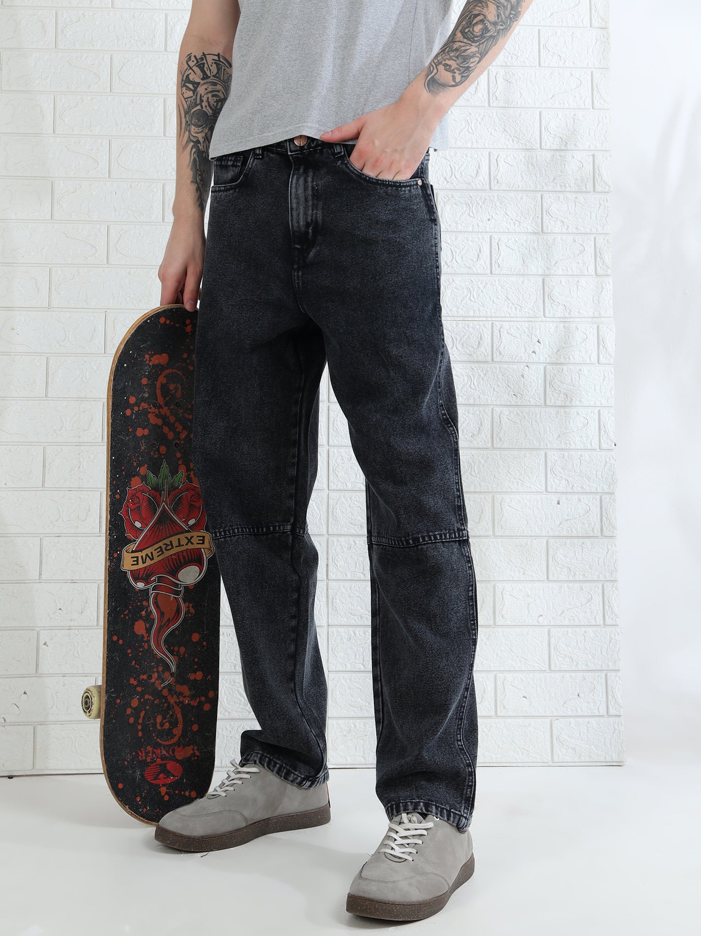 A person standing against a white brick wall, wearing a gray t-shirt, black mid-rise waist jeans, and white sneakers. They are holding a skateboard with a red design.