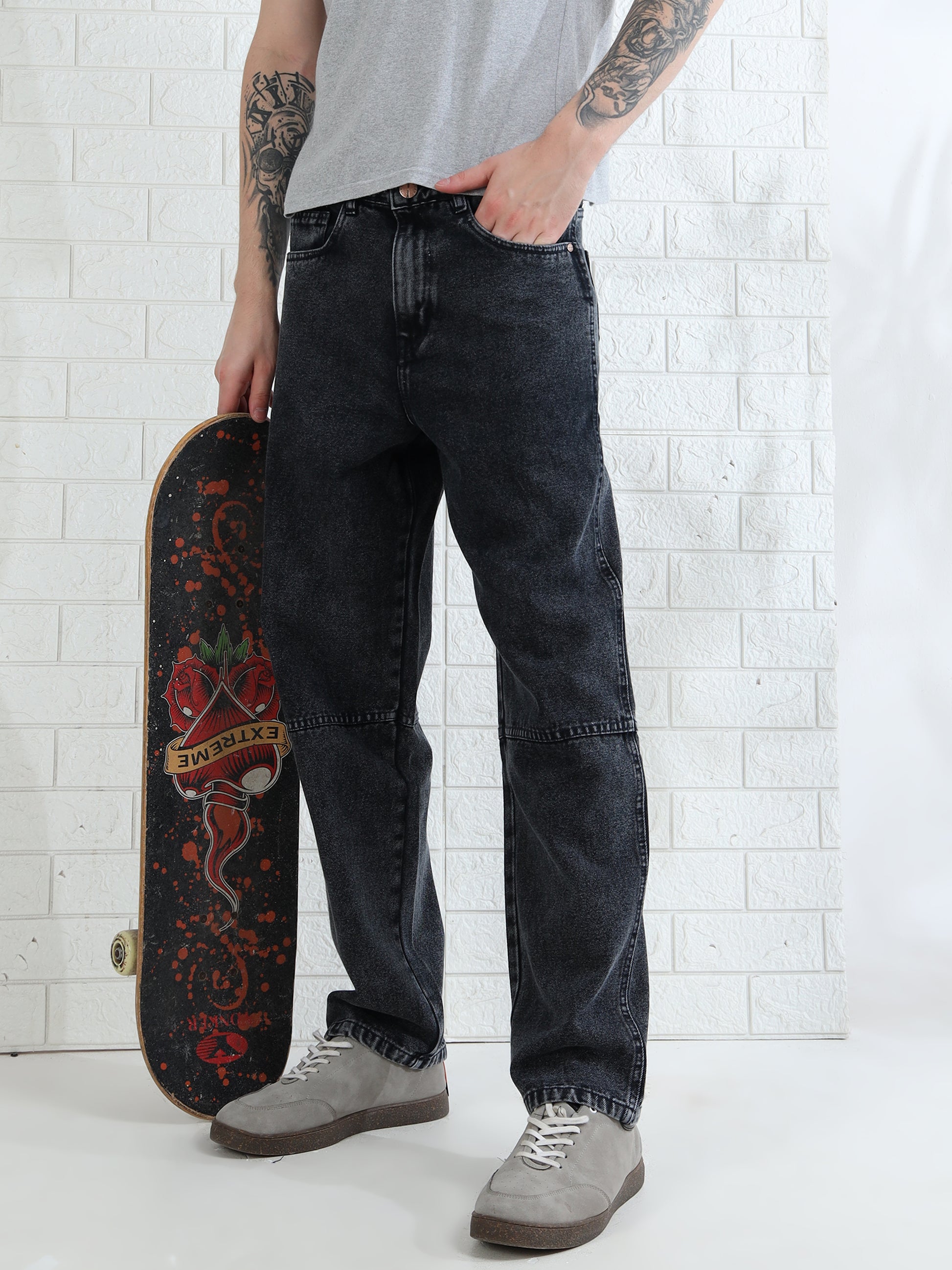 A person standing against a white brick wall, wearing a gray t-shirt, black mid-rise waist jeans, and white sneakers. They are holding a skateboard with a red design.