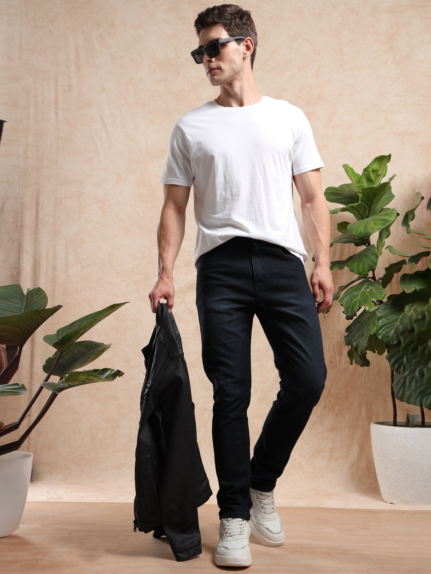 Man holding a black jacket in a minimalistic indoor setting with plants.