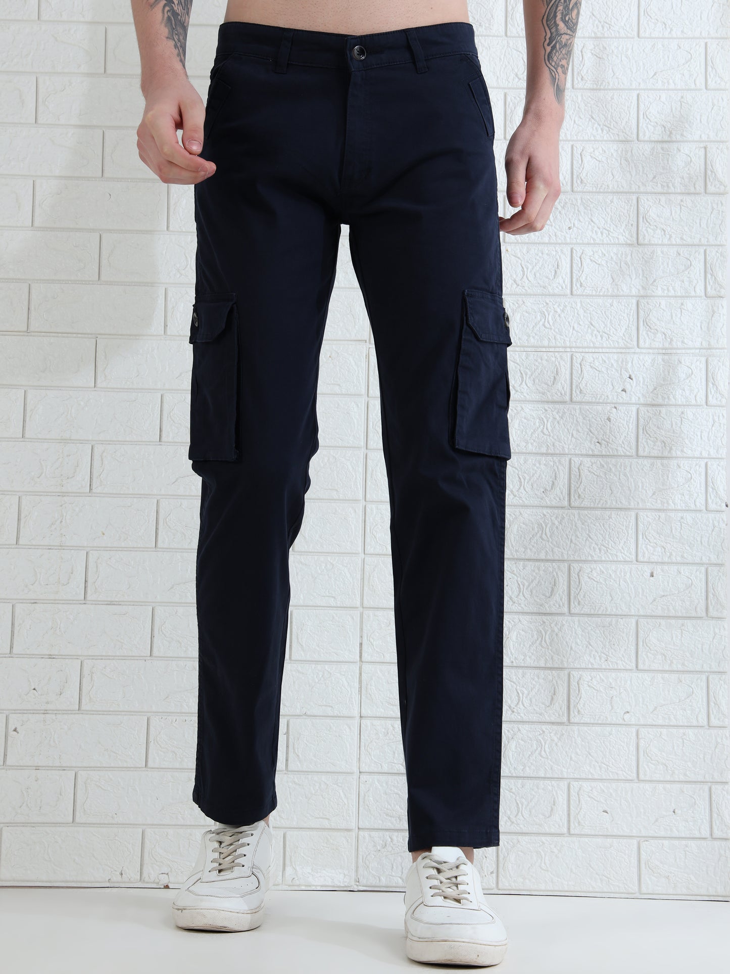 A person standing against a white brick wall wearing navy cargo pants and white sneakers.