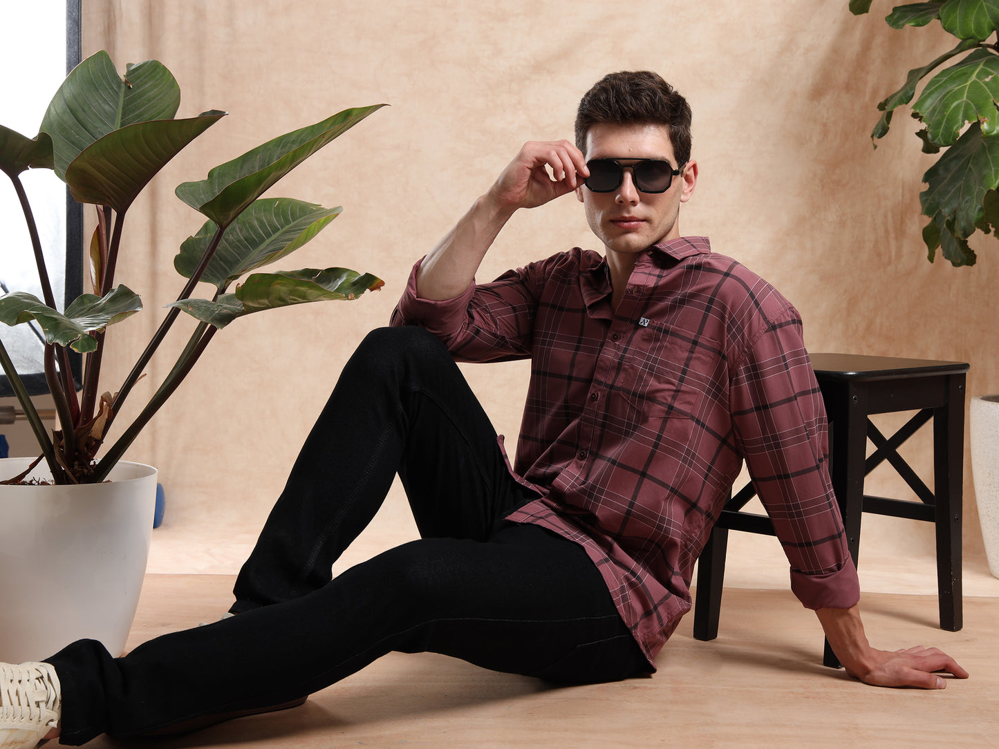 Man wearing sunglasses and a plaid shirt sitting on the floor with plants around.