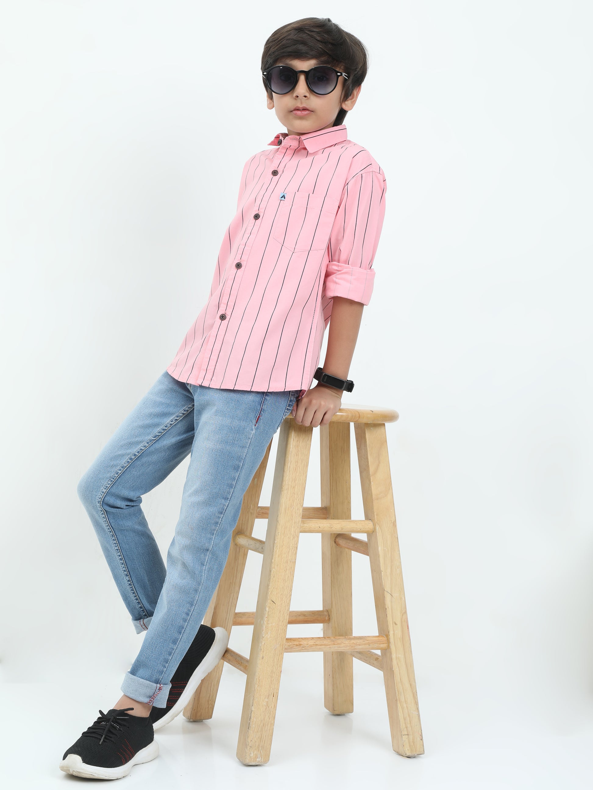 Child wearing a pink shirt and blue jeans sitting on a wooden stool against a white background