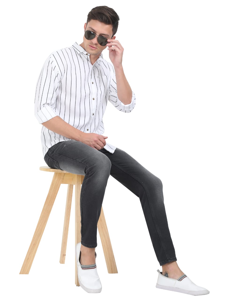 Man sitting on a stool wearing a striped shirt, black jeans, and white shoes, holding sunglasses.